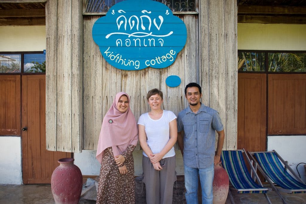 a muslim woman and a muslim man standing in front of a guest house with a foreign woman. they smile at the camera.  