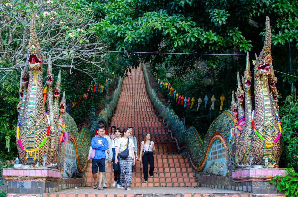 Wejście do świątyni Doi Suthep, widok ze schodów. Grupa turystów schodzi przy głowach smoków. 
